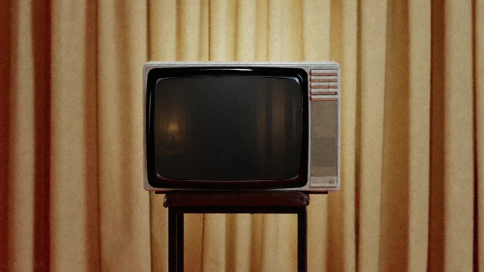 An unlit vintage television on a wooden stand, in front of warmly lit gold curtains.